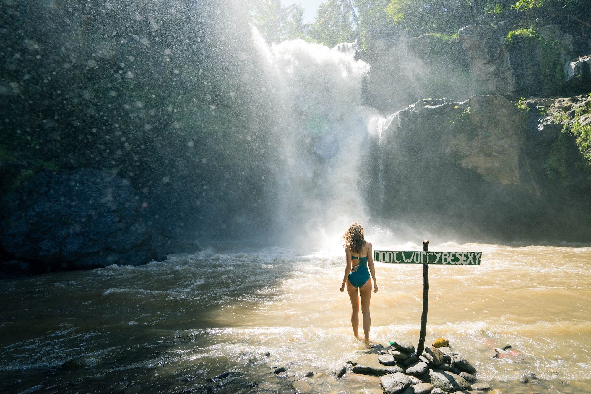 A person stands by a river in front of a majestic waterfall, sunlight filtering through the mist, with a wooden signpost nearby.
