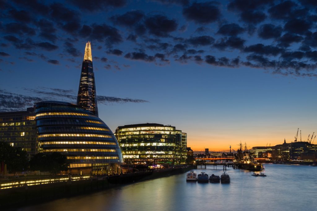This image features the London skyline at dusk, highlighting the illuminated Shard skyscraper, with the Thames River and buildings along its banks under a cloudy sky.