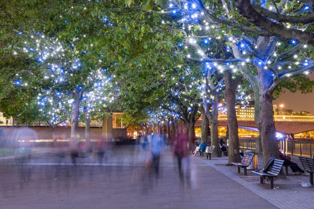 Trees adorned with blue lights line a promenade at dusk, with blurred figures of people walking and benches on the side, creating a vibrant, urban evening scene.