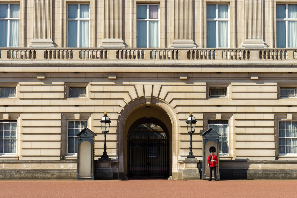 The image shows a person standing guard in traditional red and black attire outside a large, ornate arched entrance to a classic sandstone building.