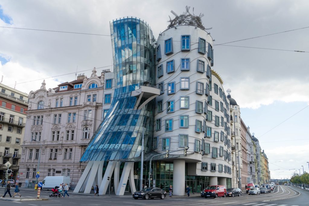 An unconventional building with a curved glass facade stands out among traditional architecture on a city street with cars and pedestrians under a cloudy sky.