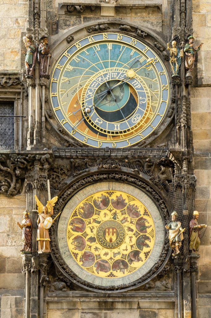 The image shows the Prague Astronomical Clock with astronomical dial, zodiacal ring, and four figures flanking the clocks on a decorated stone facade.
