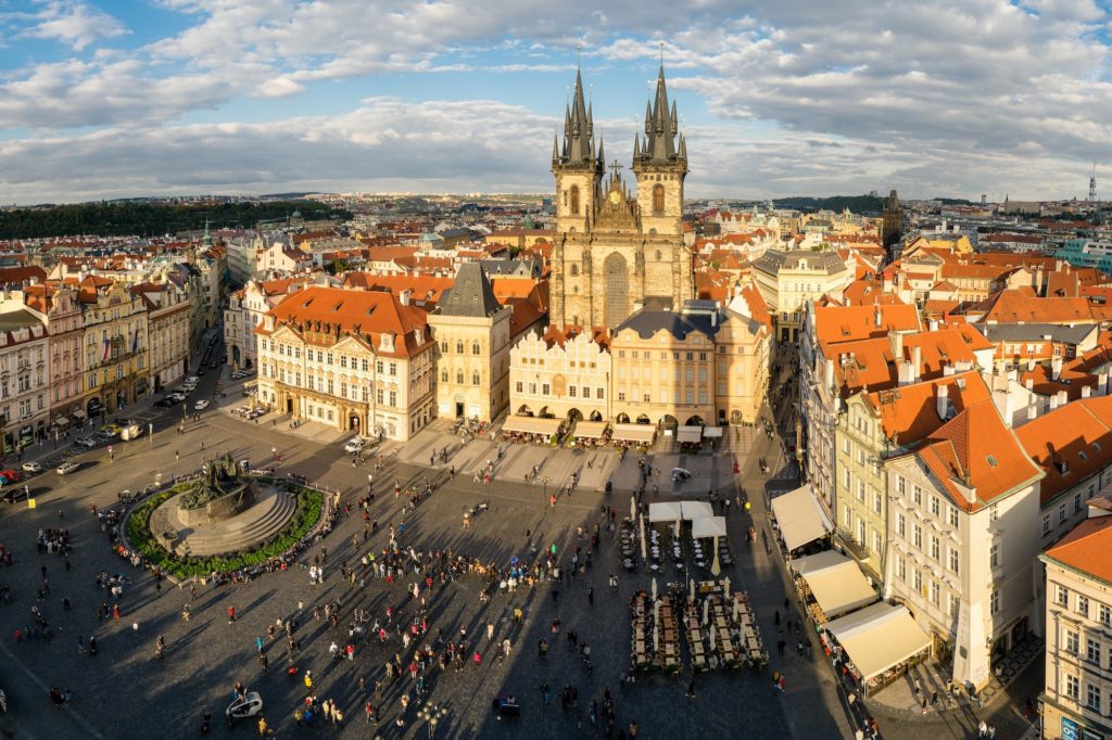 Aerial view of a historic European square with a prominent Gothic church, ornate buildings, and people scattered around, under a partly cloudy sky.