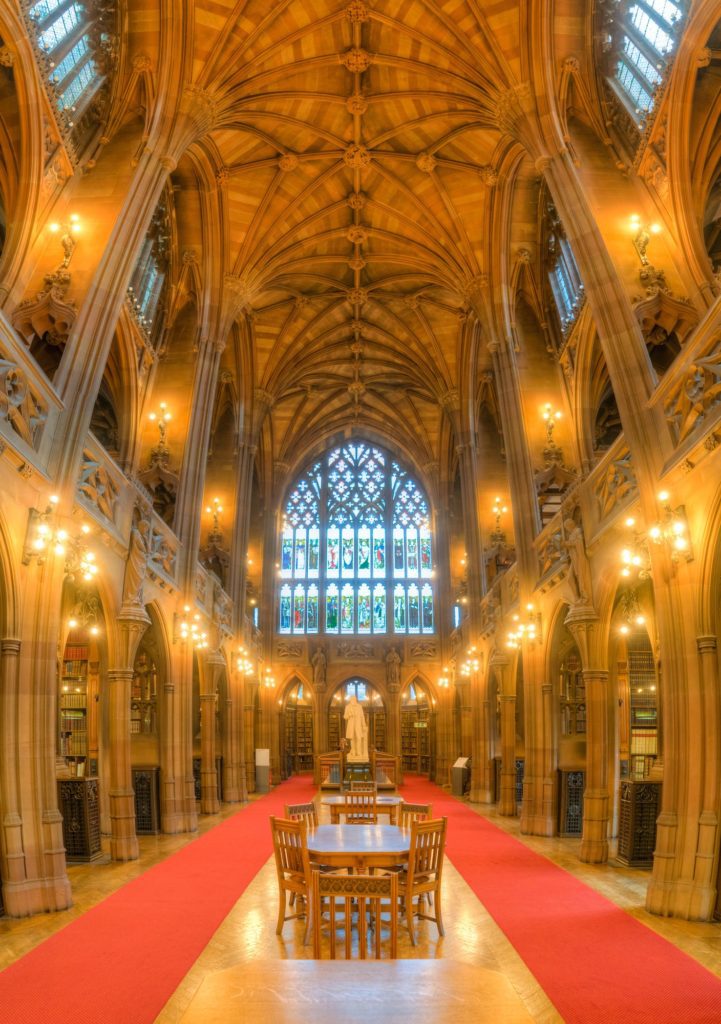 This image shows an elaborate gothic-style interior with vaulted ceilings, wooden bookshelves, a red carpet, and a large stained-glass window in the background.