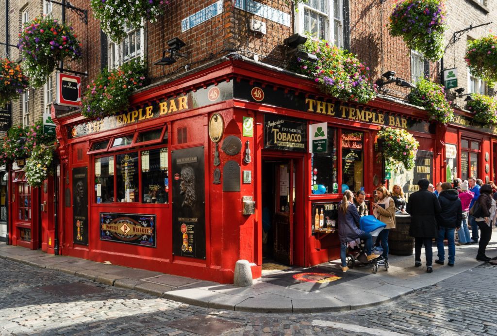This image shows a vibrant red traditional Irish pub, The Temple Bar, adorned with greenery, with people seated outside and others walking by on cobblestone streets.