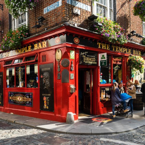 This image shows a vibrant red traditional Irish pub, The Temple Bar, adorned with greenery, with people seated outside and others walking by on cobblestone streets.