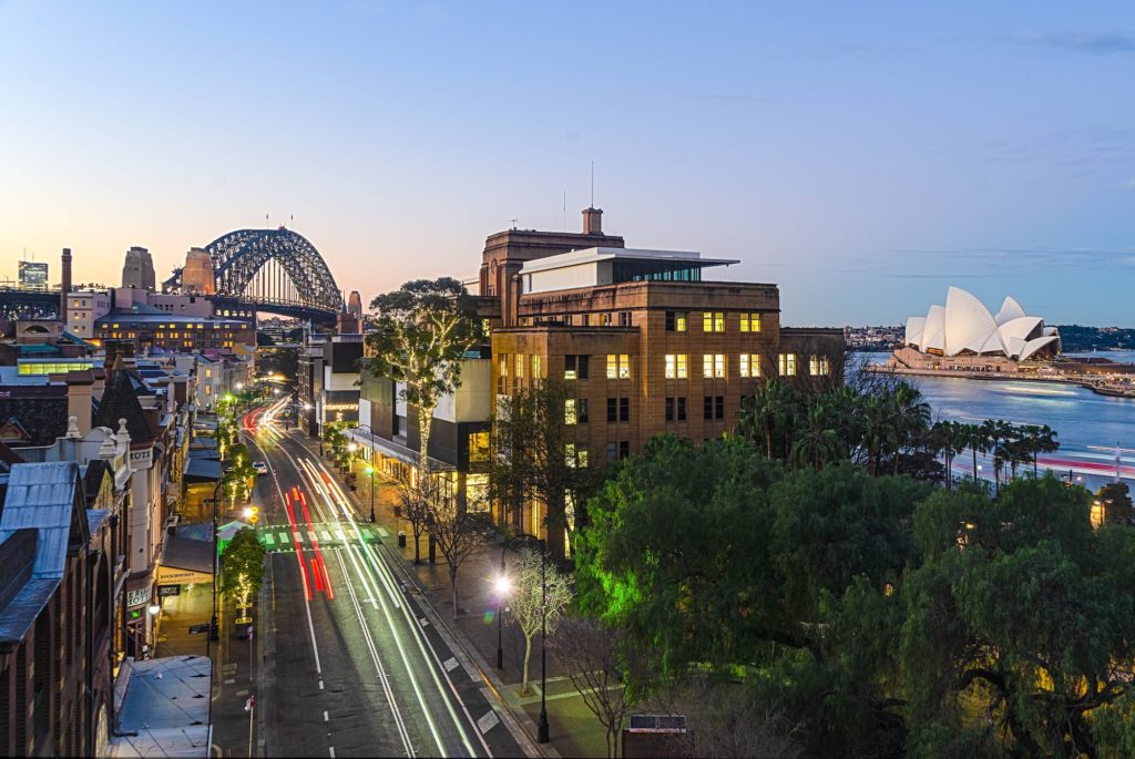 This image captures a cityscape at twilight with the iconic Sydney Harbour Bridge and Sydney Opera House, alongside illuminated streets and moving vehicles.