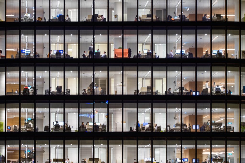 The image captures a multi-story office building at night with numerous illuminated windows, showing people working at desks and walking around inside.