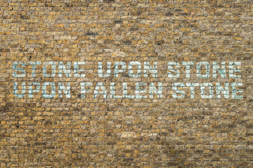 The image shows a weathered brick wall with the phrase "STONE UPON STONE UPON FALLEN STONE" painted or stenciled in blue letters.