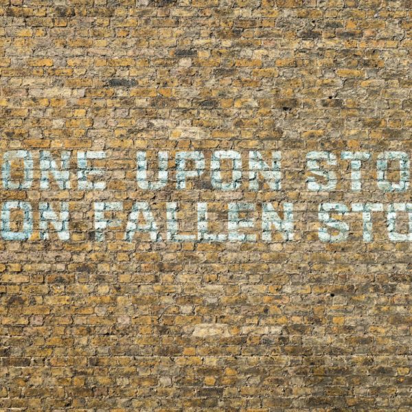 The image shows a weathered brick wall with the phrase "STONE UPON STONE UPON FALLEN STONE" painted or stenciled in blue letters.