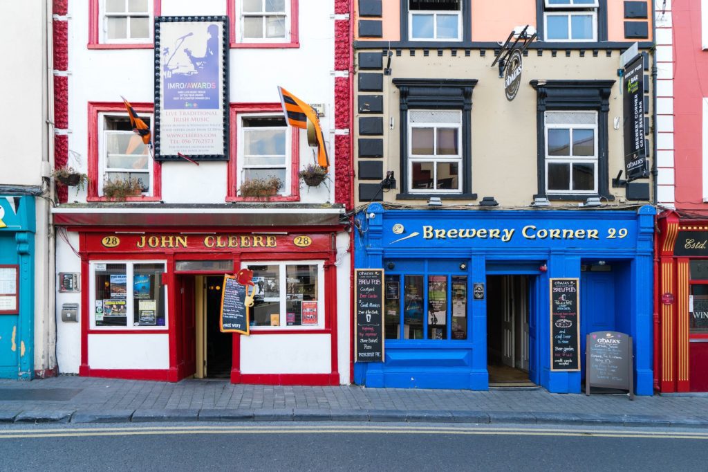 Two colorful storefronts side by side, one red and one blue, with signs indicating a pub and a brewery, located on a city street.