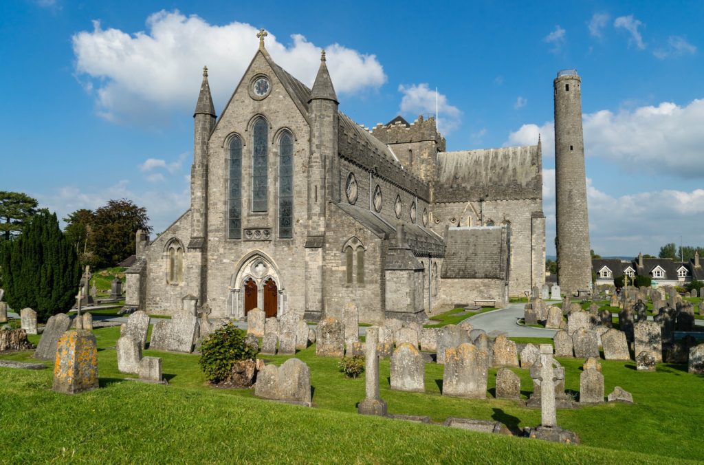 A historical stone church with a prominent tower stands beside a cemetery with numerous gravestones. It's a clear day with few clouds in the sky.