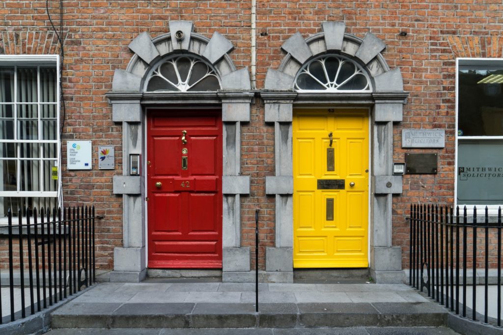 Two vibrant, contrasting colored doors—red and yellow—stand side by side in a traditional brick building facade with symmetrical stone arches and steps.