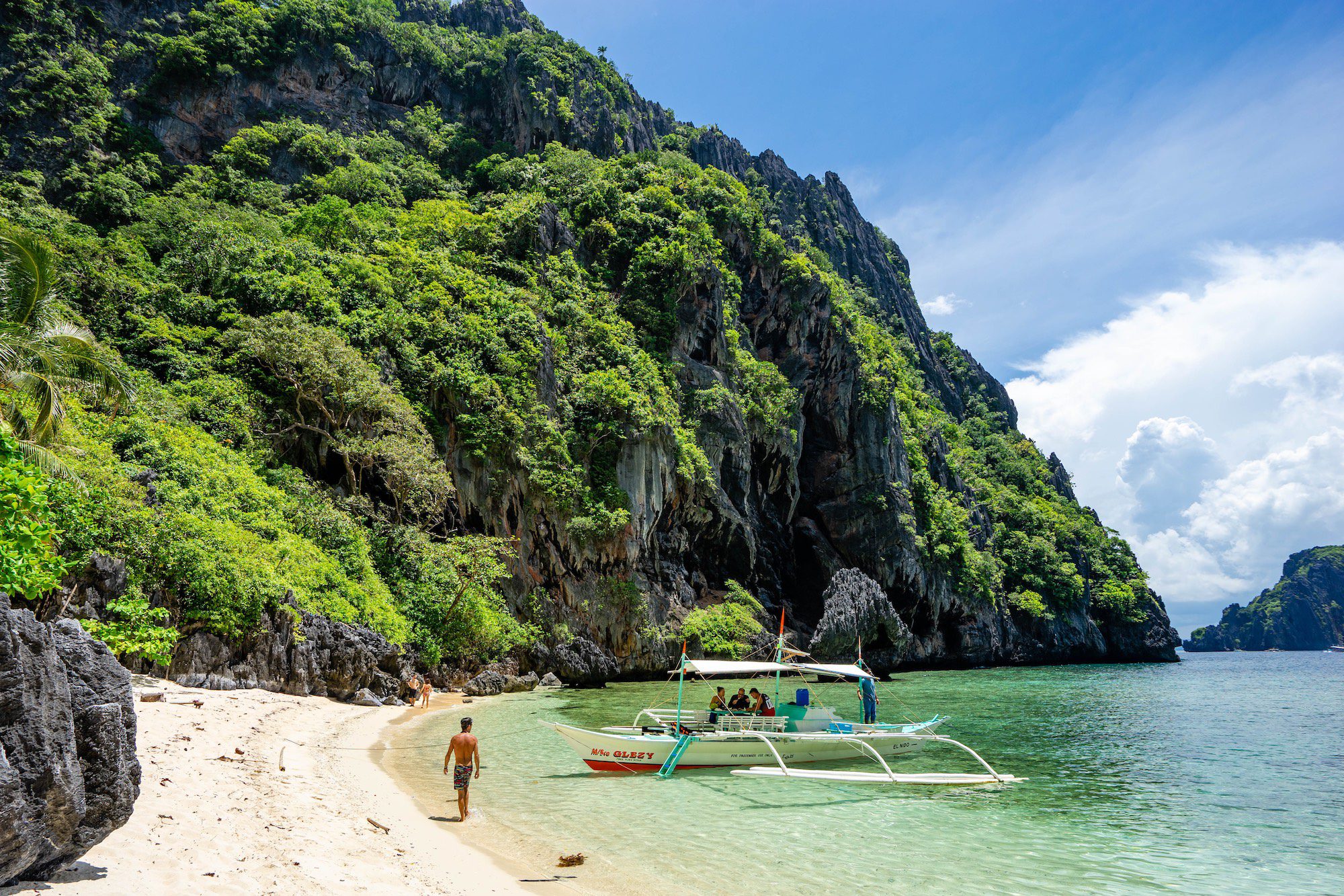 A tropical beach with clear blue skies, a limestone cliff, lush foliage, a traditional boat, and a person walking on the white sand.