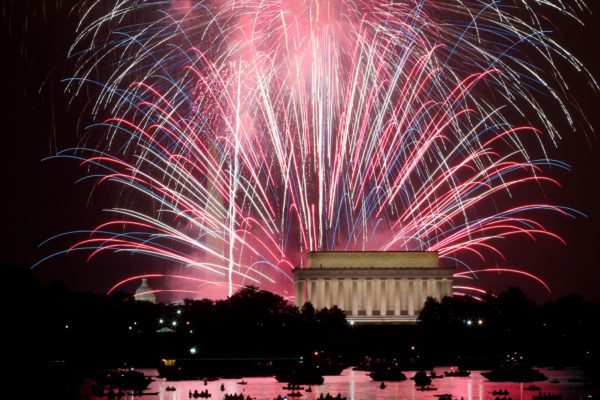A vibrant fireworks display illuminates the night sky above the Lincoln Memorial. Reflections of the fireworks are visible on the water's surface.