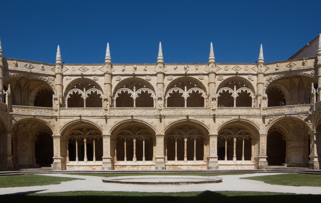 This is a Gothic-style cloister with ornate, arched colonnades, pointed arches, and a well-manicured central lawn; clear blue skies are visible above.