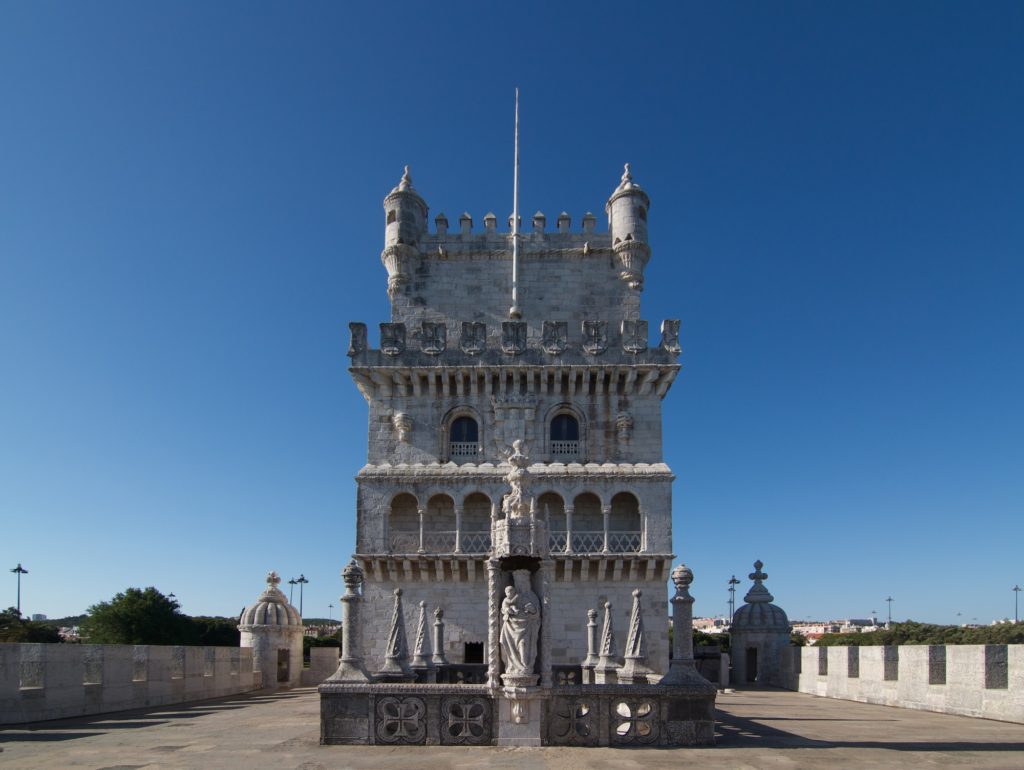 An ornate historical tower with Gothic and Manueline architectural elements stands under a clear blue sky, featuring intricate stone carvings and turrets.