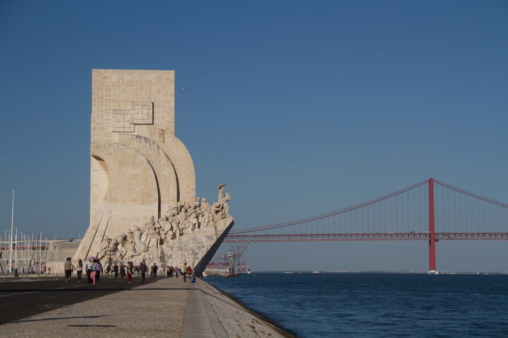 This image features the Monument to the Discoveries in Lisbon, Portugal, with people strolling nearby and the 25 de Abril Bridge in the background.