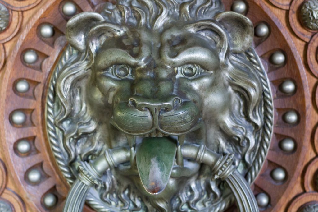 This image depicts an ornate lion head door knocker made of metal, set against a wooden door with decorative studs, exhibiting classical design elements.