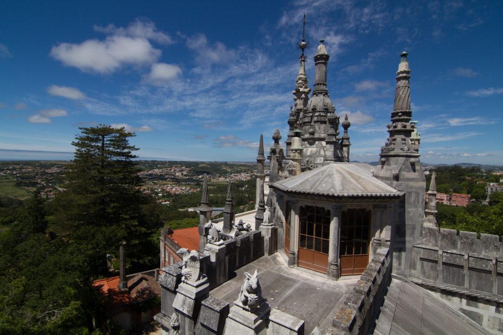 An ornate structure with spires, on a sunny day, overlooks a lush landscape. Blue sky with clouds, architectural details, and distant views characterize the scene.