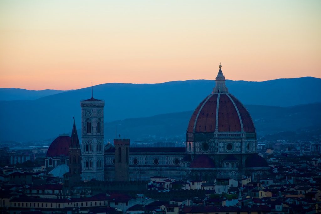 The image shows the Florence Cathedral with its iconic red dome silhouetted against a sunset, with blue to orange gradient skies and distant mountains.