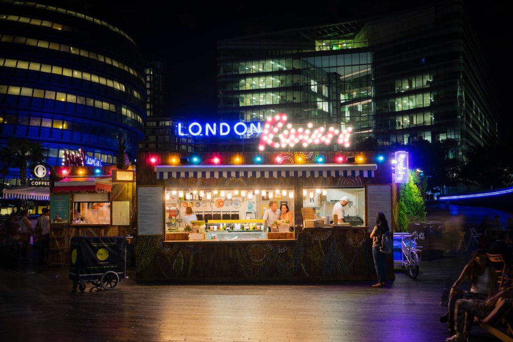 A vibrant outdoor food stall at night with "London Burger" in neon lights. People are attending the stall, surrounded by modern buildings and soft illumination.