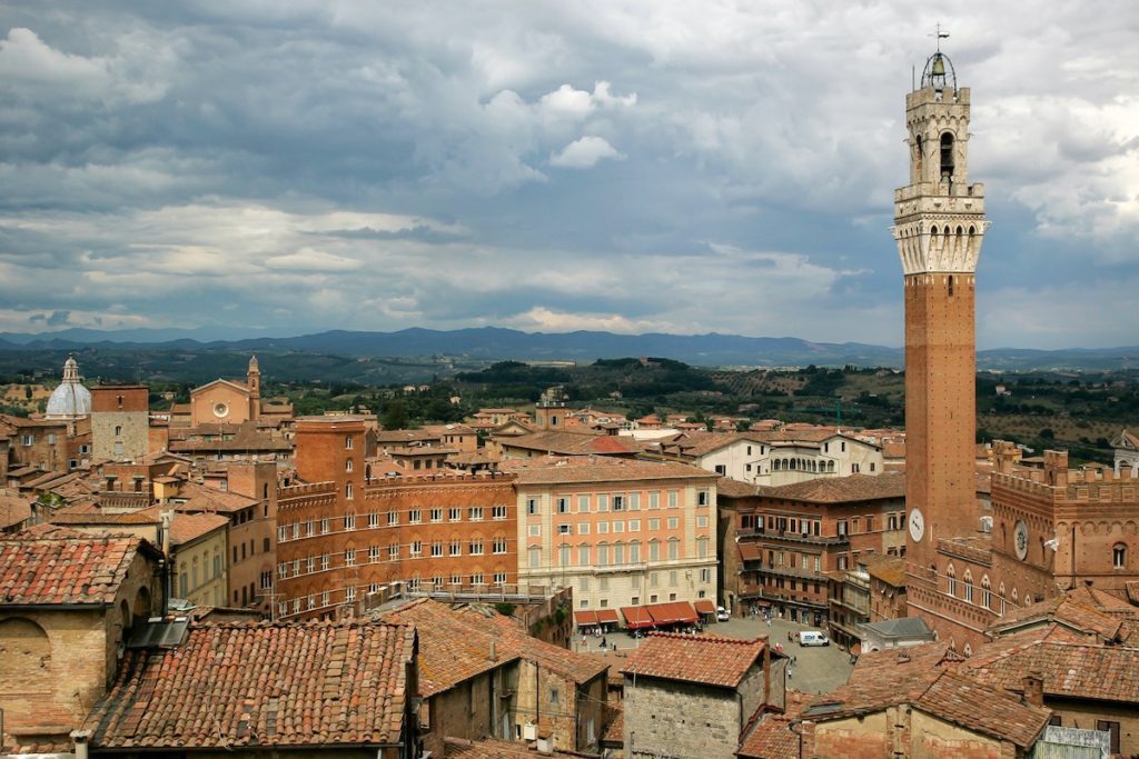An old European town with terracotta rooftops, featuring a prominent bell tower. Cloudy skies loom overhead, suggesting a large, historic, picturesque setting.