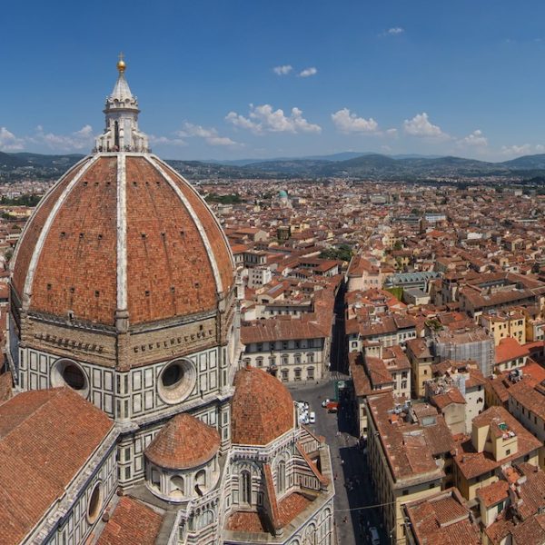 An aerial view of Florence's iconic Duomo with its large dome and surrounding terracotta-roofed buildings, set against a backdrop of distant hills and blue sky.