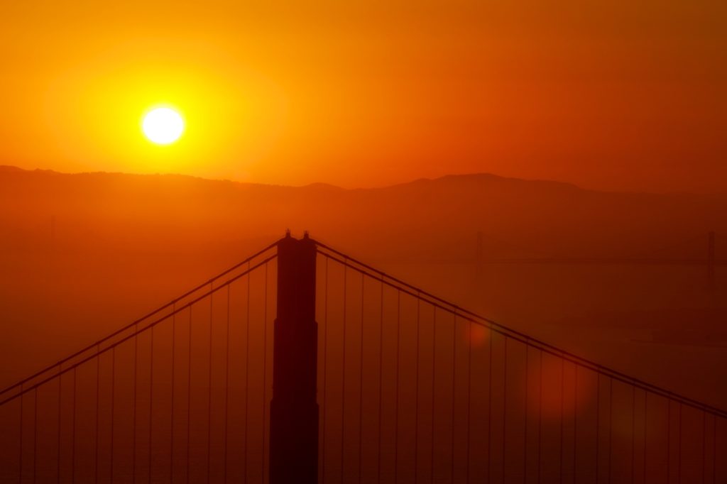 A breathtaking sunset with golden hues, featuring the silhouette of a suspension bridge tower and cables against a softly illuminated sky and distant hills.