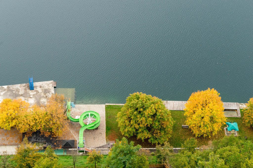 Aerial view of a waterfront with a textured green slide, trees with autumn leaves, a building rooftop, and a calm blue water body adjacent to it.