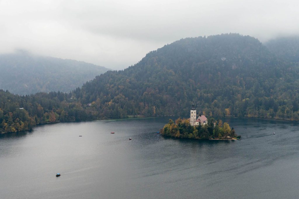 An aerial view of a misty lake with a small island hosting a white building, surrounded by forested hills in a tranquil, overcast setting.
