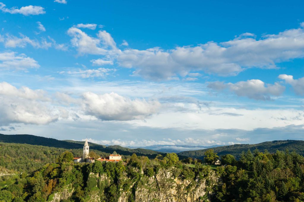 A scenic view showcases a church with a prominent steeple perched atop a rocky outcrop, surrounded by lush greenery under a partly cloudy sky.