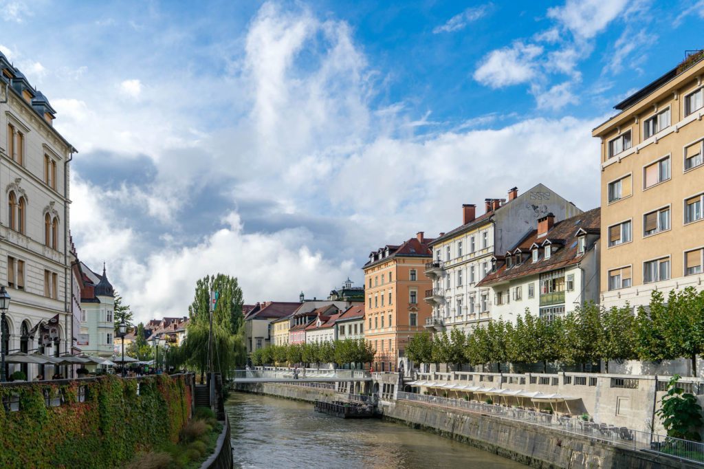 A picturesque European cityscape with historic buildings lining a canal, under a partly cloudy sky. Greenery adorns the water's edge, reflecting a serene environment.