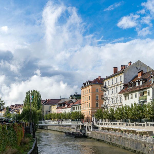 A picturesque European cityscape with historic buildings lining a canal, under a partly cloudy sky. Greenery adorns the water's edge, reflecting a serene environment.