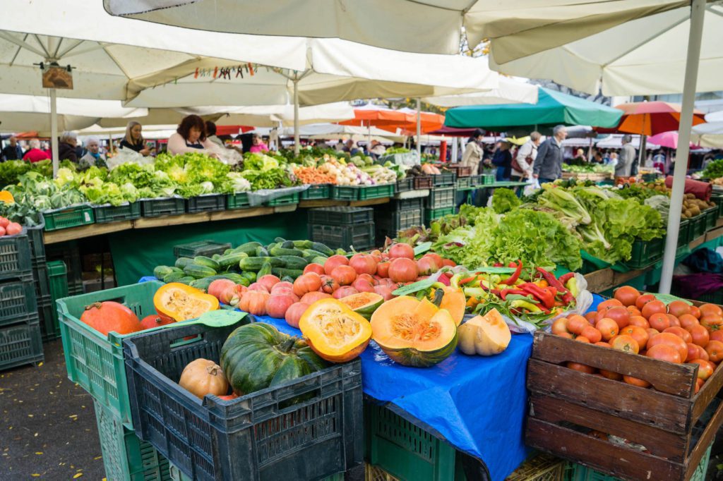 An outdoor market with various fresh vegetables and fruits on display, including leafy greens and tomatoes, with people browsing under white canopies.