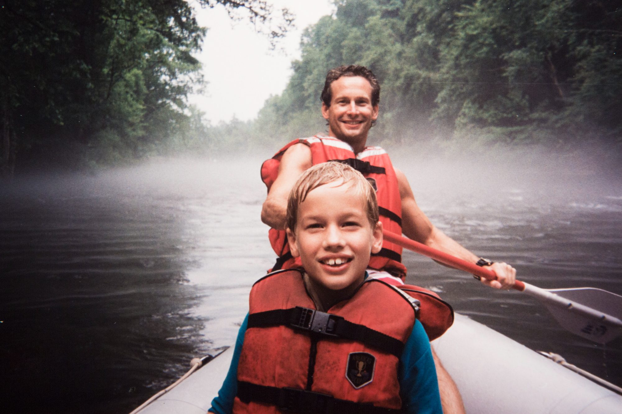 A person and a child wearing life jackets are smiling in a canoe surrounded by mist on a tranquil river, suggesting a joyful outdoor adventure.