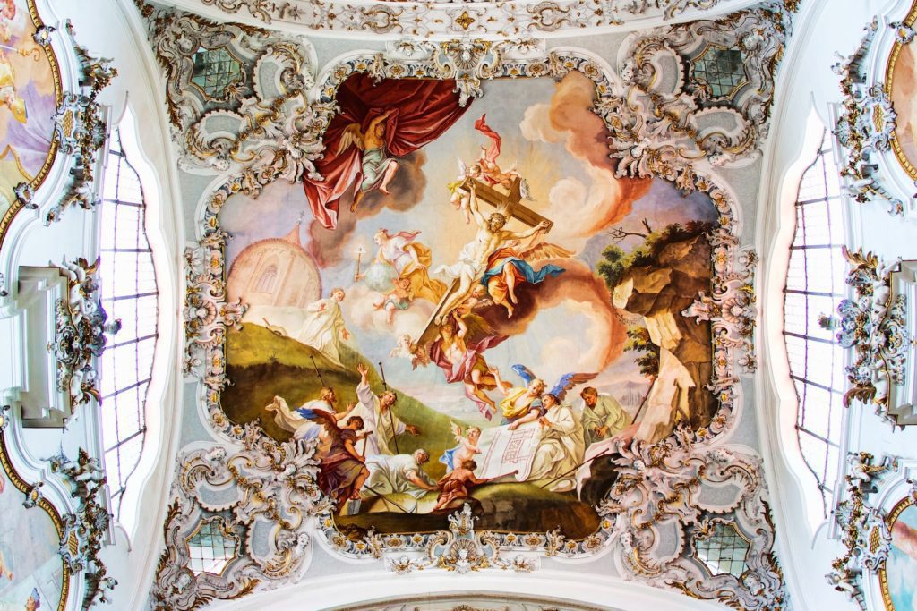 This image showcases a vibrant, baroque ceiling fresco with dynamic figures, possibly depicting a historical or religious scene, framed by elaborate stucco ornamentation.