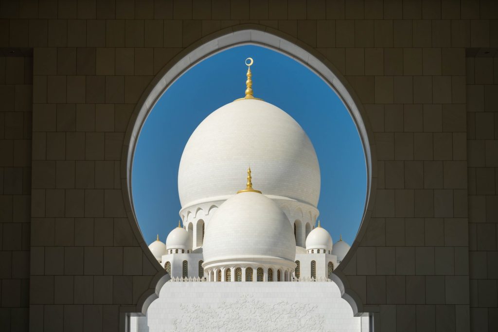 This image frames a white domed structure with gold accents through an archway, showcasing Islamic architectural design under a clear blue sky.