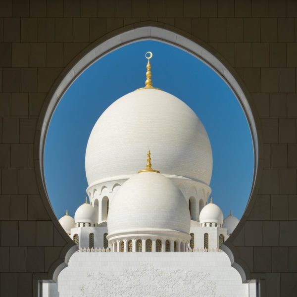 This image frames a white domed structure with gold accents through an archway, showcasing Islamic architectural design under a clear blue sky.