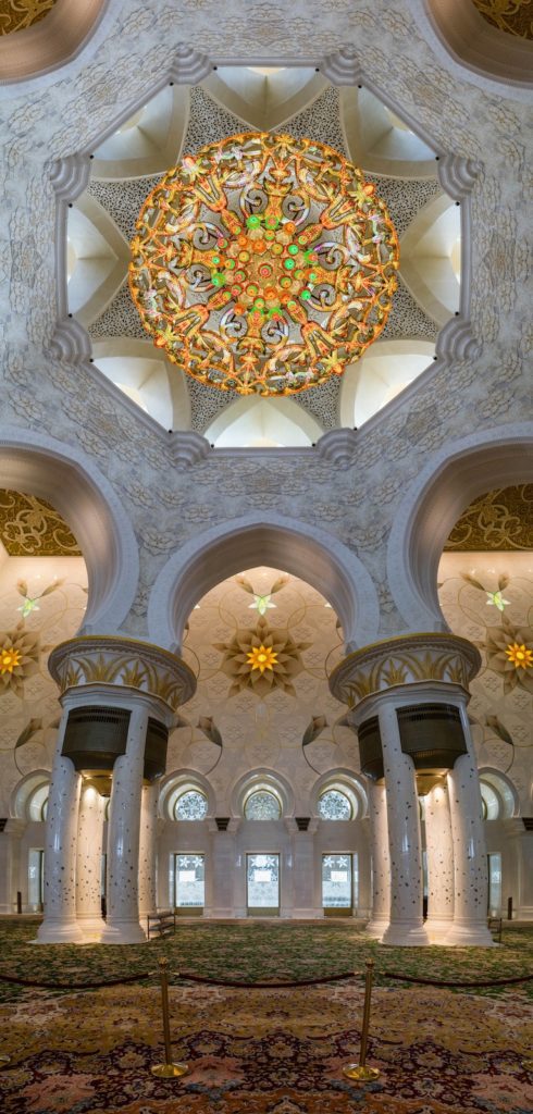 An ornate interior with elaborate floral patterned domes, arches, and columns, featuring a large, intricately decorated chandelier in a grand, symmetrical space.
