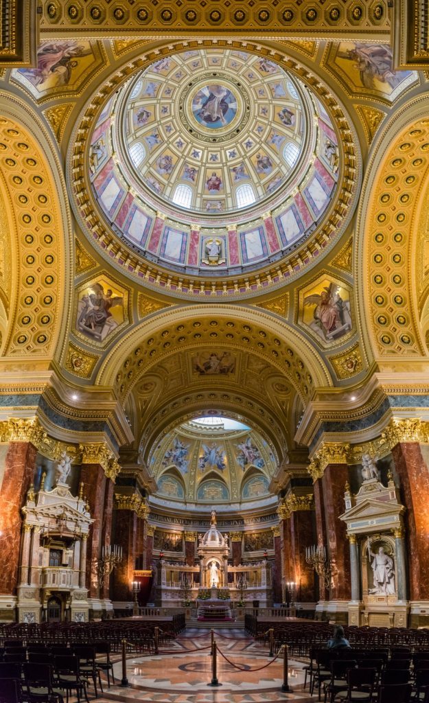 An opulent church interior with a grand dome, intricate frescoes, golden embellishments, marble columns, an altar, and rows of wooden pews.
