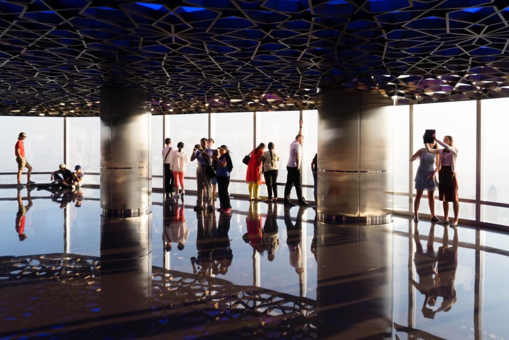 Indoor observation deck with reflective floor, several people standing and taking photos against vast windows, bright light, geometric ceiling patterns, and a modern ambiance.