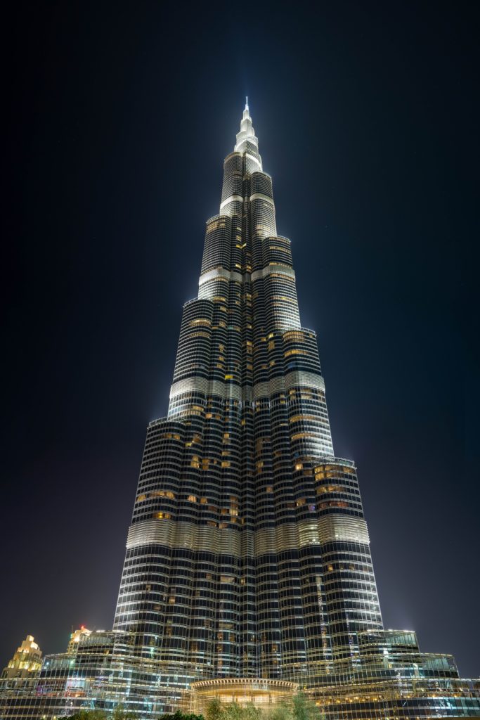 The image shows the illuminated Burj Khalifa at night, towering against a dark sky, with its distinct silhouette and layered architecture highlighted.
