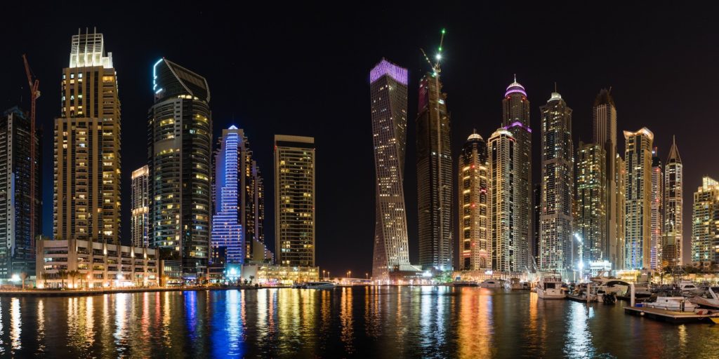 This image captures a night skyline of a modern city with illuminated skyscrapers reflecting on the water, alongside docked boats under a clear sky.