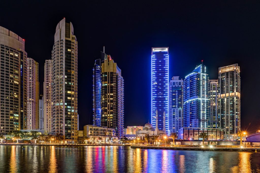 This is a night scene of a modern skyline with illuminated skyscrapers reflecting on a calm water surface, showcasing urban architecture and city lights.