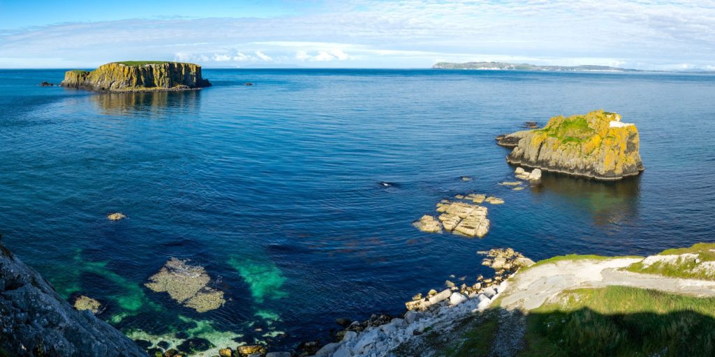 A panoramic view of a calm sea with several small, rugged islands covered in patches of greenery, and clear waters revealing underwater rocks.