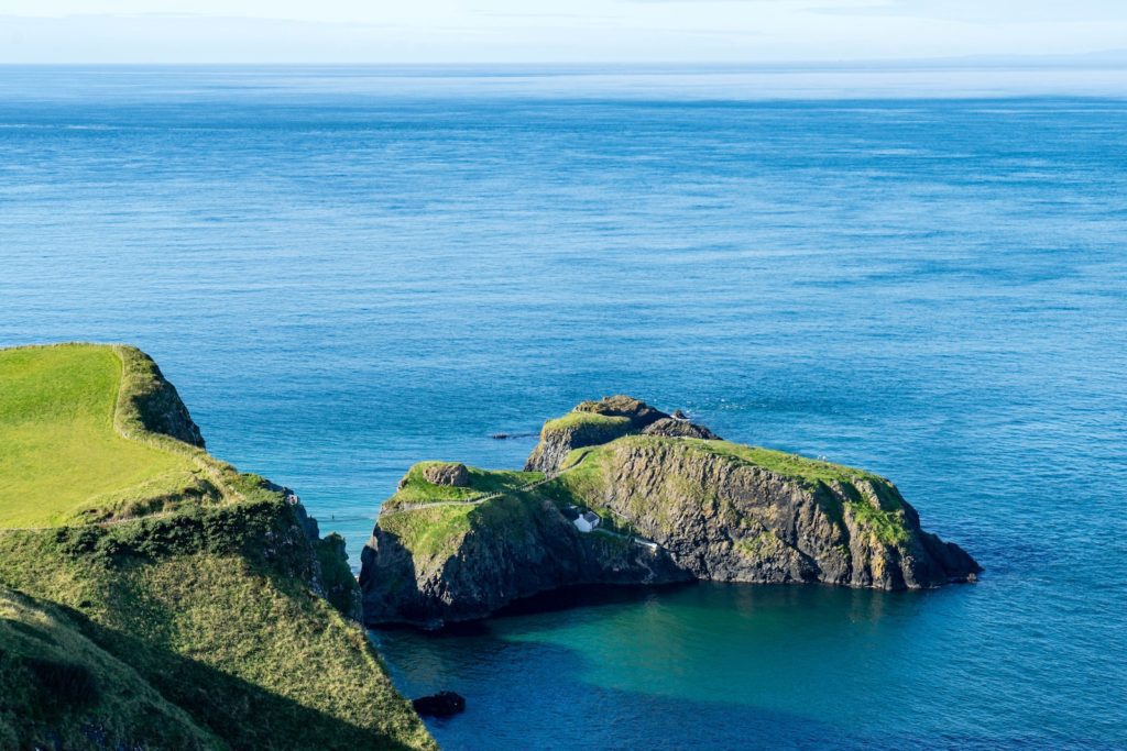 A beautiful coastal scenery showing lush green cliffs overlooking a serene blue sea under a clear sky. The landscape appears calm and undisturbed.