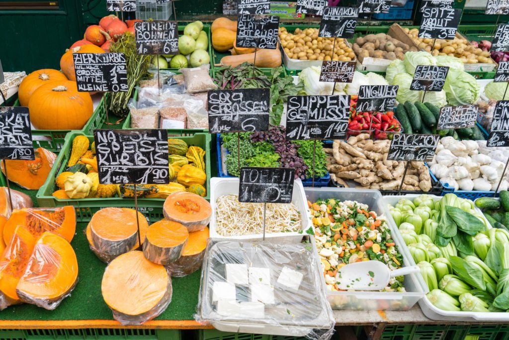 The image shows a vibrant array of fresh vegetables and food items on display at a market stall, with prices clearly labeled on black signs.