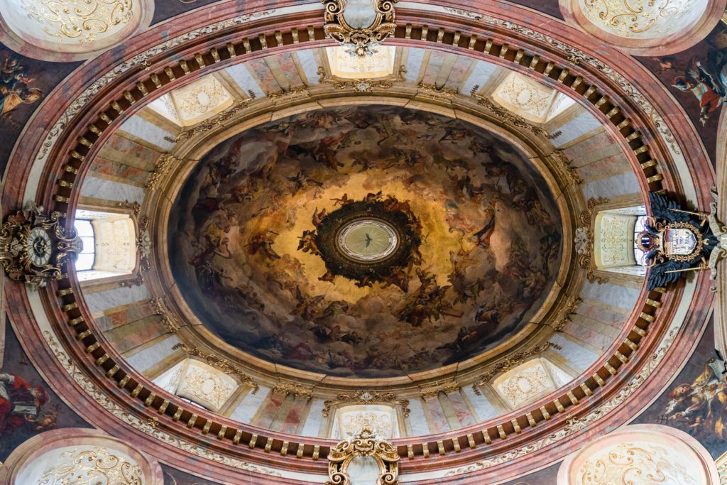The image shows the ornate interior dome of a grand building, featuring intricate frescoes, opulent chandeliers, and decorative architectural details.