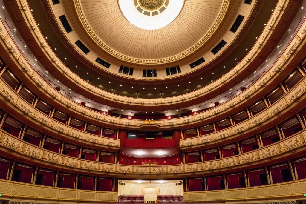 A grand theater interior with multiple ornate balconies, red seats, a decorative ceiling, and an empty stage. It appears opulent and traditional in design.